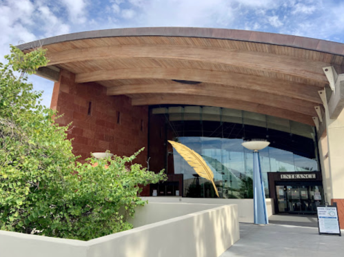 Modern building entrance with a curved wooden roof and large glass windows, surrounded by greenery.